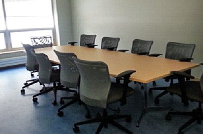 9 chairs surround a boardroom-style table. Sun shines through a large window behind them.