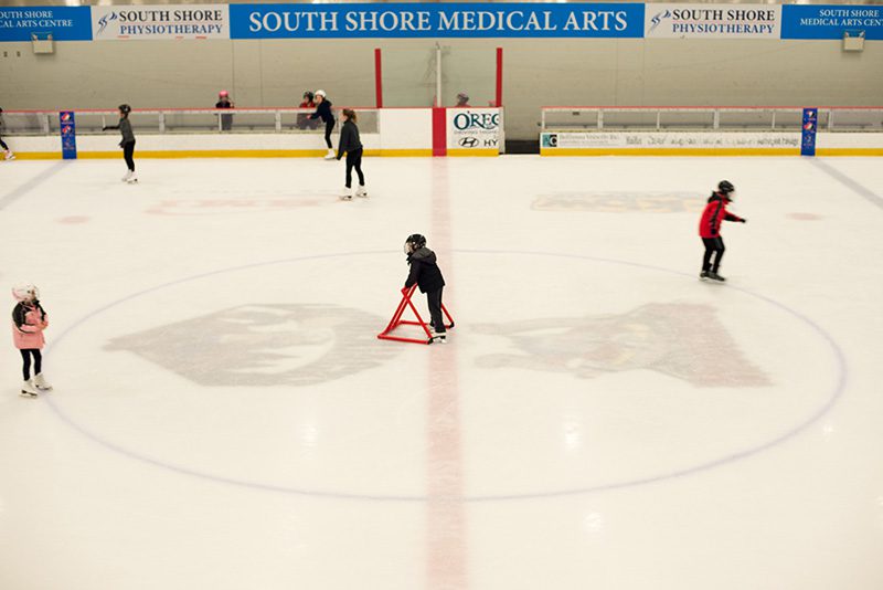 A few children skate on the LCLC arena ice. A young child pushes a bright red skating aid.