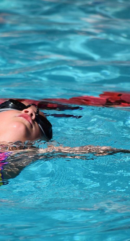 A girl wearing goggles floats on her back in a swimming pool.
