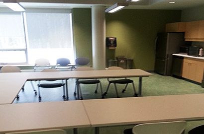A long row of classroom-style desks with chairs. A small kitchen with a large fridge lines the wall. A large window lets in bright sunlight.
